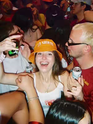 Cover image shows a crowded scene at an outdoor event, likely a tailgate or party. In the center, a woman with long brown hair, wearing an orange bucket hat that says LA SIENTA, smiles widely with her mouth open. She's wearing a white tank top. Many people surround her, some holding drinks or seemingly engaged in conversation. The background suggests a bustling, energetic environment with people in various outfits, some in sunglasses, and what appears to be the University of Southern California (USC) context is implied in the surrounding.