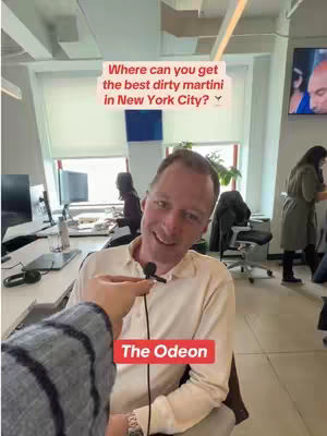 A man with short hair and a light-colored shirt is holding a microphone in an office setting. Above him is a text overlay question which asks: "Where can you get the best dirty martini in New York City?" A red tag below the microphone reads "The Odeon".