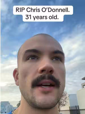 A close-up shot of a man with a bald head and a thick mustache, looking upwards with a concerned expression against a cloudy sky backdrop. Text overlayed on top reads "RIP Chris O'Donnell. 31 years old."