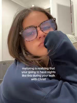 A selfie-style shot features a young woman with short brown hair and eyeglasses, gazing slightly downward with a subdued expression. She is wearing a dark blue hoodie, with her hand partially covering her mouth. The background appears to be a neutral-toned interior. Overlay text towards the bottom describes a moment of personal reflection.