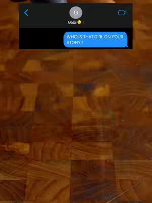 The image shows a screenshot of a phone screen displaying a text message conversation. The background appears to be a wooden surface. The main element is a message bubble that says, "WHO IS THAT GIRL ON YOUR STORY!!" The screen imitates an iPhone display, and there are interface elements at the top, including a back arrow, a contact icon labeled "Gabi," and a video call button. The color scheme is dark with the message bubble standing out with its blue color and a white font on top.