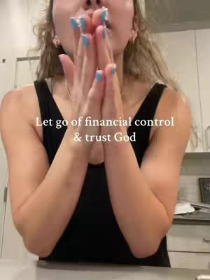 A person, likely a woman, is seen from the chest up. She's holding her hands up in front of her face, with fingers touching as if in prayer or offering. Her nails are painted a bright blue. The background is a blurred interior with neutral colors. The text overlay reads 'Let go of financial control & trust God'.