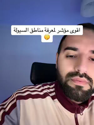 A man wearing a tracksuit and posing for the camera, in front of a blue background. Above his head is a white rectangle with arabic text on it, and a yellow emoji.