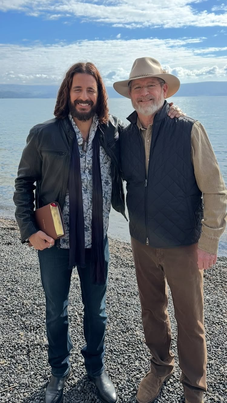 Two men are standing outdoors near a body of water, possibly a lake, on a cloudy day. The man on the left is wearing a black leather jacket, a patterned shirt, dark pants, and holding a book. The man on the right is wearing a hat, a vest, and a button-down shirt. They are both smiling at the camera.