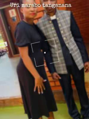 A medium shot shows a woman and a man posing closely together. The woman is on the left, wearing a black dress. The man on the right is wearing a blazer-like jacket with a plaid pattern. The background appears to be a building exterior.