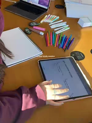 The cover image shows a top-down view of a table cluttered with study materials. A laptop is open in the background, showing a screen with a website visible. A notebook, pens, and highlighters are organized on the table alongside a tablet displaying a webpage. A hand is holding the tablet in the foreground. The color palette is warm, with the table surface a light wood tone and the items adding pops of color. The layout is arranged to create an impression of studious preparation.