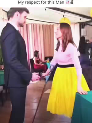 A man in a suit is standing in front of a woman in a yellow skirt and pink top. They appear to be in a professional setting. A third person is seated at a table in the background
