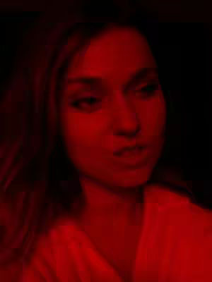 A close-up shot of a woman's face illuminated in red light against a dark background. Her expression is subtle, with a slight pursing of the lips. The lighting creates high contrast, drawing attention to her features.