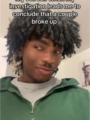 A selfie-style shot featuring a young Black man with curly, dark hair. He wears a green zip-up jacket over a gray t-shirt. The background is a simple indoor setting. The man has a slight smirk and is looking to the side, suggesting a knowing or mischievous expression. The overall composition is a close-up, focusing on his face and upper body.