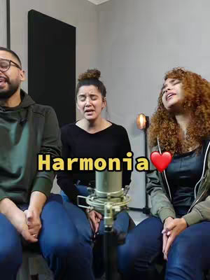 The cover image shows three people singing into a microphone in a studio setting. The person on the left is a man with glasses, wearing a dark green hoodie, and the person on the right is a woman with curly red hair, wearing a black tank top and a green jacket. The person in the middle is a woman with a bun, wearing a black top. The text "Harmonia" appears in yellow, with a red heart emoji.