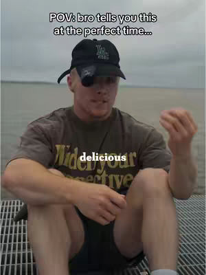 A man is sitting outdoors, dressed in a brown t-shirt and black shorts, with a black cap on. The background shows a body of water and a cloudy sky, with the text overlaying the image.
