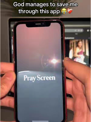 The image shows a person holding a smartphone displaying a specific app screen. The phone is held in front of a blurred background. The app screen has the words “Pray Screen” in white text. The background is dark, possibly an outside scene with what appears to be a glimpse of a woman on another screen blurred in the background.