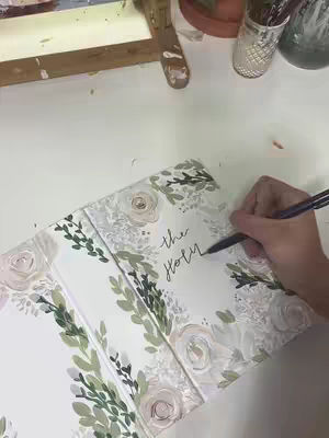 A person is writing the words "the holy" on the open pages of a bible that is decorated with floral designs. The bible is open on a white surface with other art supplies visible in the background.
