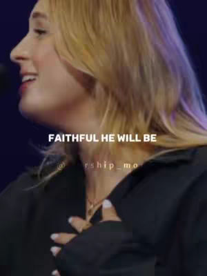 A close-up shot of a woman's profile while singing, with overlayed text. She is wearing a black top and has blonde hair. The background is dark, and the lighting seems to be from a spotlight, creating shadows on her face. Overlayed text in white font says "FAITHFUL HE WILL BE".