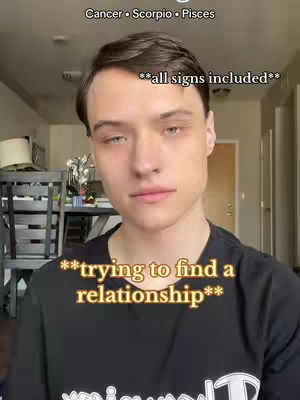 A selfie-style shot featuring a fair-skinned person with short brown hair looking directly at the camera. Text overlays include zodiac signs and phrases referencing relationships. The background suggests an indoor setting.