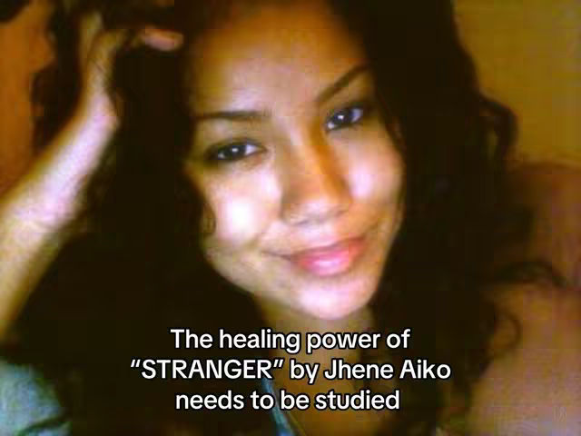 A close-up shot of a woman with dark, wavy hair smiling at the camera. The background is out of focus and appears to be a light color. Text overlayed on the image discusses Jhene Aiko's song 'Stranger'.