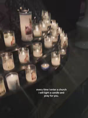 The image shows a display of lit candles in glass holders on a tiered metal stand, likely within a church or similar setting. A large pillar candle is visible at the back, and smaller candles with printed images around the glass, are arranged in a circular manner. The color palette is mainly dark, with soft, warm light emanating from the candles.