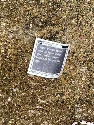 A close-up shot of a crumpled piece of paper with a printed message lying on a textured, gravel-like surface. The paper is slightly tilted, and the background appears to be a mix of small pebbles and possibly partially wet or reflective patches. The paper seems to be from a news or memo-style document, with the beginning of the message legible. The lighting is natural, and the color palette is dominated by earth tones.