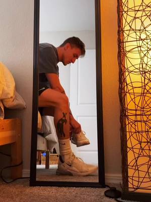 A man is captured in a mirror, tying his shoe. He is wearing a gray t-shirt, shorts, and white sneakers. A tattoo is visible on his leg.