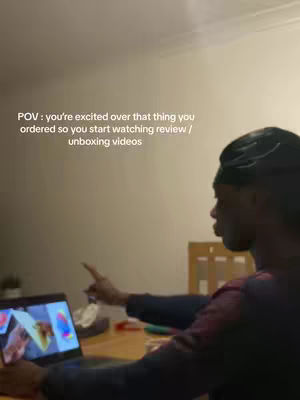A man is seated at a wooden table, looking at a laptop screen. The scene is shot from a medium angle, highlighting the man's profile as he points towards something on the screen. The background is neutral, with a plain wall and a glimpse of a chair, possibly indoors. The laptop screen displays colorful images, suggesting the man is watching a video.