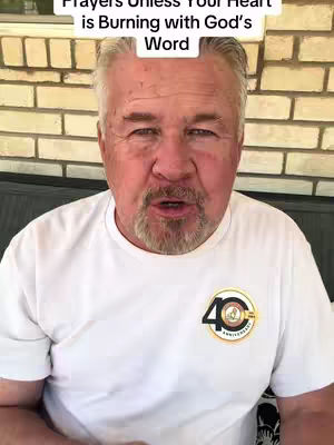 A man with gray hair and a beard is centered in the frame, looking directly at the viewer. He is wearing a white t-shirt with a circular emblem on the chest and is positioned in front of a light-colored brick wall. Above his head, text overlays the image.