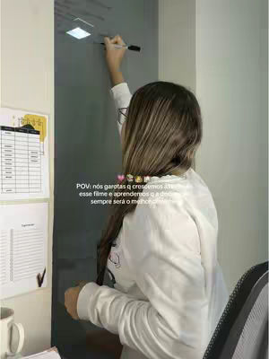A woman with long brown hair is shown from the back, wearing a white long-sleeved shirt. She is standing in front of a glass surface, possibly a whiteboard or window, and writing on it with a black marker. To her left, there are some papers and a mug on a desk. The scene appears to be an office or a workspace. The color palette includes whites, grays, and a touch of yellow.