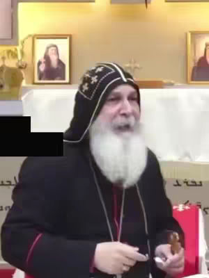 A man wearing a black and patterned head covering with a long white beard is speaking. He is positioned in front of a wall and some objects. Behind him there are several framed pictures on the wall.