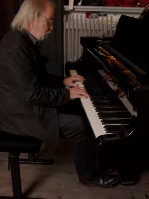 An older man with long white hair and glasses plays a black piano in a dimly lit room. He is seated on a piano bench, and his hands are on the keys. The image is composed with a focus on the musician and the instrument, with the background slightly out of focus.