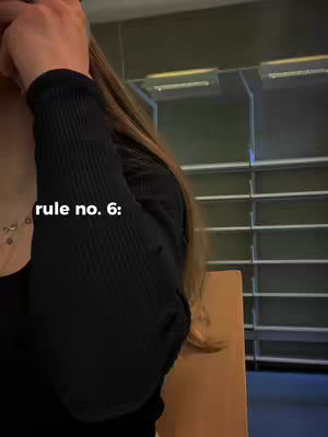 A person with long brown hair partially obscures their face, with their left arm and the sleeve of a black shirt visible. The background features a blurred interior with rows of lights and a wooden chair partially in view.