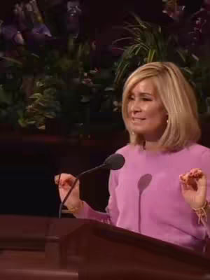 A woman with blonde hair is speaking at a podium. She's wearing a pink shirt and the background is a blurred image of greenery. A microphone is in front of her.