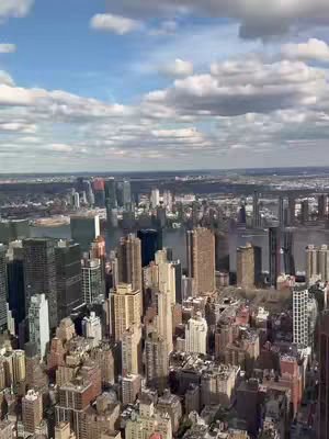 An eye-level shot from above showcases a dense cityscape under a bright, slightly overcast blue sky. Tall skyscrapers of various heights and architectural styles fill the frame. The foreground is packed with buildings, while the background reveals a skyline of distant structures and a stretch of water. White and grey clouds softly diffuse the sunlight. The overall composition emphasizes the vastness and density of the urban environment.