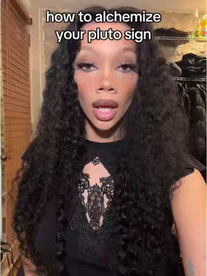 A woman with long, dark wavy hair is centered in the frame, looking directly at the camera with a slight open-mouth expression. She wears a black top with a decorative lace pattern at the chest. The background includes glimpses of a room, some shelves, and a glimpse of what looks like a hanging rack with clothes. The overlay text at the top reads, 'how to alchemize your pluto sign'.