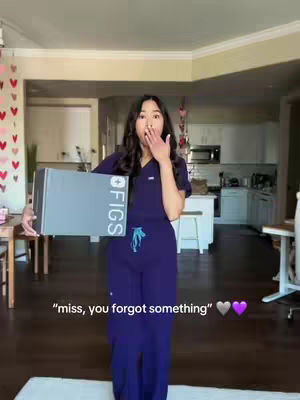 A woman stands in a home, holding a gray box labeled "FIGS." She wears a purple scrubs top and pants, with a surprised expression and her hand covering her mouth. The background shows a living room and kitchen.