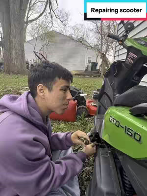A young man, with a dirty face, is crouched down and working on a bright green scooter. He is wearing a purple hoodie. The background shows a grassy area, a white house, parked lawnmowers, and a large tree.