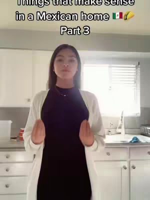 A young woman stands in a kitchen, looking towards the camera. She's wearing a dark dress and a white cardigan. The background shows kitchen cabinets, a window, and other kitchen items.