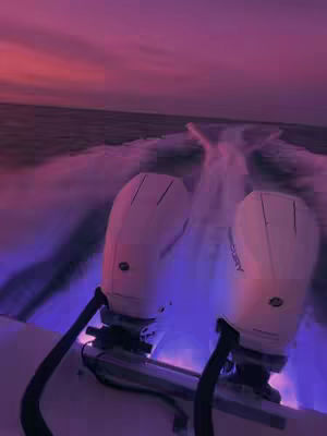 A low-angle shot from behind a boat, showcasing its white engines with their lower sections lit with purple lights. In the background, the ocean and a dramatic sunset with pink and purple hues fill the sky.