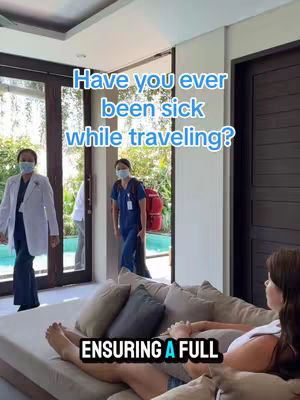 A woman lies on a beige couch in a well-lit room with a large window and glass doors. Three people wearing medical uniform are walking in the background, suggesting a medical emergency. Overlaid text asks if the viewer has ever been sick while traveling. 