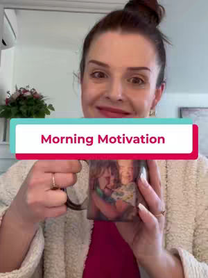 A woman with a top-bun hairstyle holds a coffee mug with a photo on it. She's wearing a white cardigan over a pink shirt, and there's a hint of a green background with a floral arrangement, adding color contrast. She smiles at the camera.