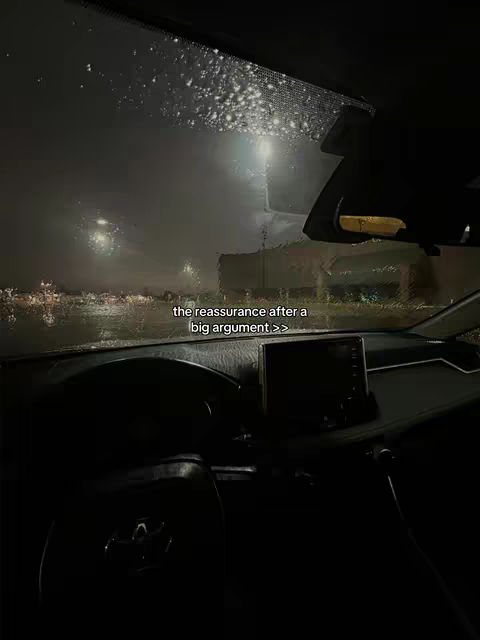 An interior car shot on a rainy night. The windshield is covered in raindrops, and the dashboard and interior are visible in the foreground. Outside, streetlights are faintly visible, and the overall mood is dark and moody.
