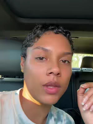 A woman with short, dark, curly hair looks directly at the camera while sitting in a vehicle. The lighting suggests the sun is shining in from the side as she sits in the interior of a car with the headrest of the seat visible behind her.