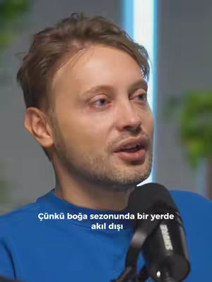 A close-up shot of a man with light brown hair and a slight beard. The man is wearing a blue shirt and appears to be speaking into a microphone, which is held in his hand. Text overlays the image at the bottom, with blurred elements in the background. The overall composition is focused on the man's face and upper body.