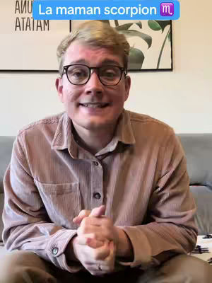 A man with glasses is seated on a gray couch indoors. He is wearing a tan corduroy shirt. Behind him is a wall with a framed art piece with a plant design.