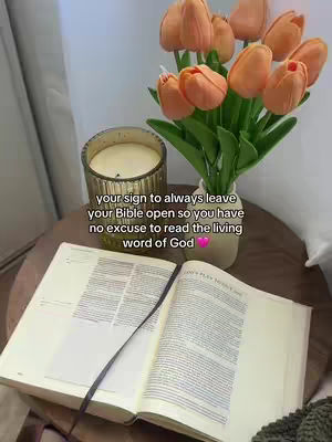 An open Bible lies on a round wooden table, with a black ribbon bookmark visible. A vase of orange tulips is in the background, along with a jar candle. The setting suggests a serene and reflective atmosphere, lit by soft, natural light.