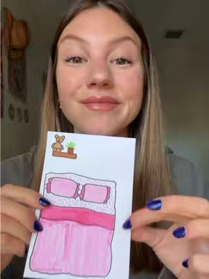 A woman holding a drawing, smiling at the camera. The drawing is of a pink bed.