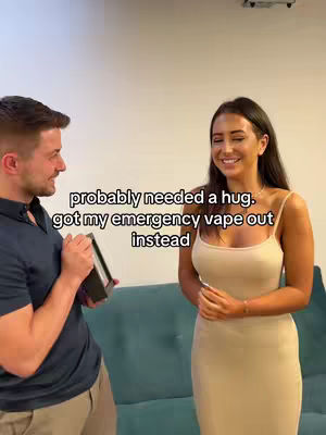A man and woman are in a room. The man is holding a box and facing the woman, who is turned toward the camera and smiling. The background includes a blue sofa and a white wall.