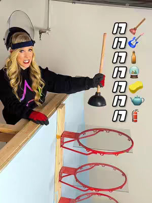 A woman in safety gear and a black hoodie stands on a wooden structure in front of a white wall with three red basketball hoops. She holds a plunger aloft, with a series of objects and related emoji icons to her right. The image is brightly lit with direct lighting.