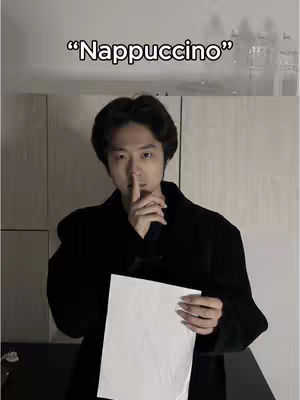 A man in a black coat holds a white paper in the foreground with his index finger touching his lips, signaling silence against a backdrop of an upper-level room. Above him is the text overlay in bold letters, "Nappuccino."