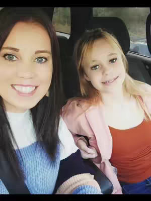 A selfie-style image featuring two smiling women seated in a vehicle. The woman on the left has dark hair and is wearing a white and blue sweater. The woman on the right has blonde hair and a pink dress over an orange top. The background shows the interior of the vehicle and a glimpse of trees through the window.