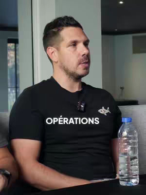 A man is sitting and talking to the camera in a well-lit indoor setting. He has short dark hair and a trimmed beard. He's wearing a black t-shirt with the white text "OPÉRATIONS" on the front, and a small logo on the left side. A water bottle sits to his right. The background is blurred, suggesting a room or interior.