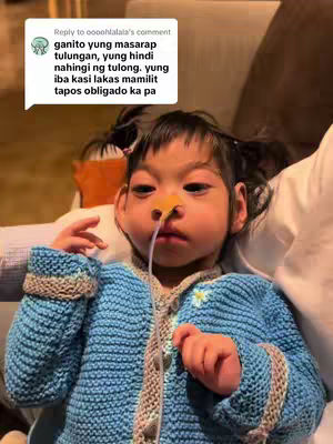 A young girl with a medical device in her nose is the subject of the image. She is wearing a blue knitted sweater with beige accents. The background is blurred, suggesting an indoor setting, possibly a bed or a couch. A chat bubble with text in a foreign language is superimposed.